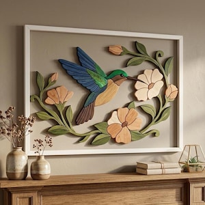 May include: A rectangular wall art piece in a white frame featuring a layered wooden hummingbird in shades of blue, green, and brown, surrounded by carved wooden flowers in light peach and green leaves, mounted on a solid light gray background.