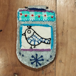 Pode incluir: Enfeite de pássaro de feltro feito à mão. O enfeite de feltro cinza apresenta um pássaro bordado em azul e branco sobre um fundo branco. A parte superior tem um padrão xadrez turquesa e vermelho. Um floco de neve azul na parte inferior.