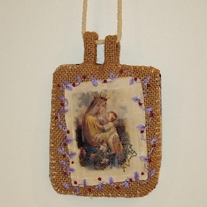 Può includere: Un ciondolo religioso rettangolare con un'immagine di Maria e Gesù, incorniciata con tela di juta e ricami viola. È appeso a una corda beige. L'immagine mostra Maria con una corona che tiene Gesù.