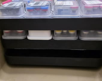 May include: A tan storage case containing a black multi-tiered organizer insert. The top tier holds four clear plastic containers with colorful lids and white labels. The middle tier contains additional clear containers, and the bottom tier is empty.