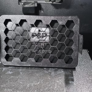 May include: A black rectangular object with a honeycomb design and the text "P&P PRINT TEC SPER" is inside a 3D printer. The object has hexagonal cutouts. The printer's interior is dark and metallic.