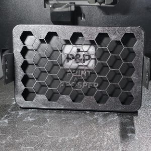 May include: A rectangular black 3D printed cover featuring a honeycomb grid pattern. The center displays the text P&P PRINT AND PROSPER. The object has small side mounting tabs with three holes each, designed for attachment to a larger mechanical device.