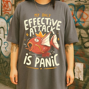 Peut inclure: T-shirt gris surdimensionné avec le texte "MY ONLY EFFECTIVE ATTACK IS PANIC" en blanc. Le graphisme représente un poisson de dessin animé rouge avec une couronne. Le t-shirt est de style décontracté et streetwear.