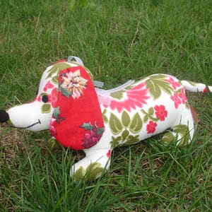 May include: A stuffed dachshund dog toy with a white body, adorned with a floral pattern in red, pink, and green. The dog has a red ear with a floral design, a black nose, and is lying on green grass.