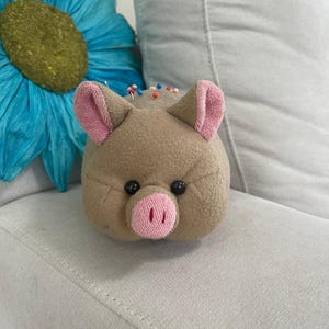 May include: A tan fabric pincushion shaped like a pig's head, with pink ears and snout, and black button eyes. Several colorful pins are inserted into the top. A large blue flower is in the background.