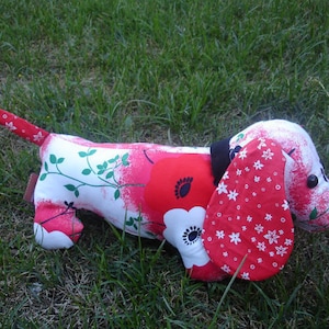 May include: A stuffed dachshund dog toy with a red, white, and green floral pattern. The dog has a red ear with white star-shaped flowers, a black nose, and black eyes. The toy is lying on green grass.