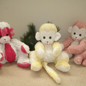 May include: Three plush monkey toys in white, pink, and yellow. The monkeys have stitched facial features and are sitting on a neutral-colored surface. One monkey is white with pink accents, another is yellow and white, and the third is pink.