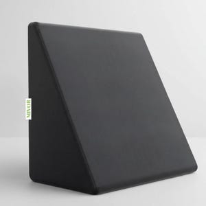 Puede incluir: Una almohada negra en forma de cuña con una superficie lisa. La almohada tiene forma triangular y está diseñada para brindar soporte. La marca "MIVOR" está impresa en una etiqueta blanca en el lateral.