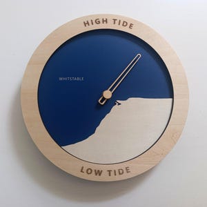 Op de afbeelding: Een ronde getijdenklok met een lichthouten frame en een diepblauwe wijzerplaat. De klok bevat de tekst HIGH TIDE bovenaan, LOW TIDE onderaan en WHITSTABLE aan de linkerkant. Een enkele lichthouten wijzer wijst naar een gestileerde witte kustlijn.