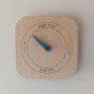 Può includere: Un orologio delle maree in legno di forma quadrata con angoli arrotondati. Il quadrante presenta un design circolare con le parole "High Tide" e "Low Tide". Una lancetta turchese indica il livello della marea. L'orologio è montato su un muro bianco.