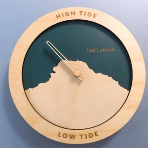 Op de afbeelding: Een ronde getijdenklok met een lichthouten frame en een donkergroenblauwe wijzerplaat. De klok heeft een lichthouten uitsnede van een kustlijn, één wijzer en de tekst HIGH TIDE bovenaan, LOW TIDE onderaan en East Lothian op de wijzerplaat.