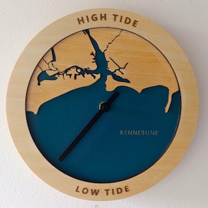 Puede incluir: Un reloj de pared redondo de madera con un mapa de Kennebunk, una sección de agua azul verdoso profundo y masas de tierra de madera clara. El texto HIGH TIDE está arriba, LOW TIDE abajo y KENNEBUNK impreso en el área azul con manecillas negras.