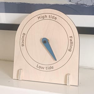 Può includere: Orologio delle maree in legno chiaro. Il quadrante riporta le scritte "High tide", "Low tide", "Rising" e "Falling". Una lancetta blu indica il livello della marea. L'orologio ha due piccoli supporti in legno.
