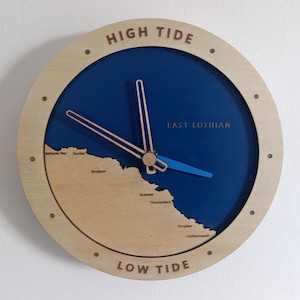 Può includere: Un orologio da parete circolare con una cornice in legno chiaro che riporta la scritta HIGH TIDE in alto e LOW TIDE in basso. Il quadrante mostra un'area oceanica blu e una mappa costiera in legno chiaro dell'East Lothian con località.