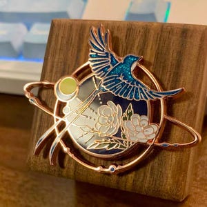 May include: A circular rose gold metal enamel pin featuring a blue glitter bird in flight above white floral blossoms. The design includes a thin orbital ring, a small yellow moon, and a dark blue background, all set within a circular frame.