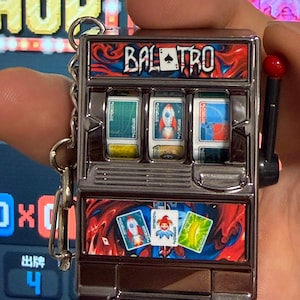 Könnte beinhalten: Ein metallischer Balatro-Spielautomat-Schlüsselanhänger mit rot-blauem Marmordesign. Die Vorderseite zeigt den Text Balatro, drei Walzen mit Kartensymbolen, einen seitlichen Hebel mit rotem Knopf und ein unteres Feld mit drei Spielkarten.