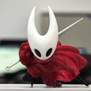 Op de afbeelding: Een 3D-geprint beeldje van een personage met een witte, hoornachtige hoofdtooi en zwarte oogopeningen. Het figuur draagt een vloeiende rode cape en houdt een zilveren naaldachtig wapen vast. Het beeldje staat in een actiepose.
