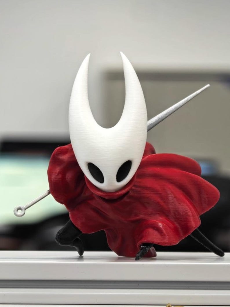 Op de afbeelding: Een 3D-geprint beeldje van een personage met een witte, hoornachtige hoofdtooi en zwarte oogopeningen. Het figuur draagt een vloeiende rode cape en houdt een zilveren naaldachtig wapen vast. Het beeldje staat in een actiepose.
