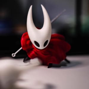 Op de afbeelding: Een beeldje van een personage uit de videogame Hollow Knight. De figuur heeft een witte kop met twee gebogen hoorns en zwarte ogen. Het draagt een rode cape en houdt een zilveren naald vast. De figuur staat op een wit oppervlak.