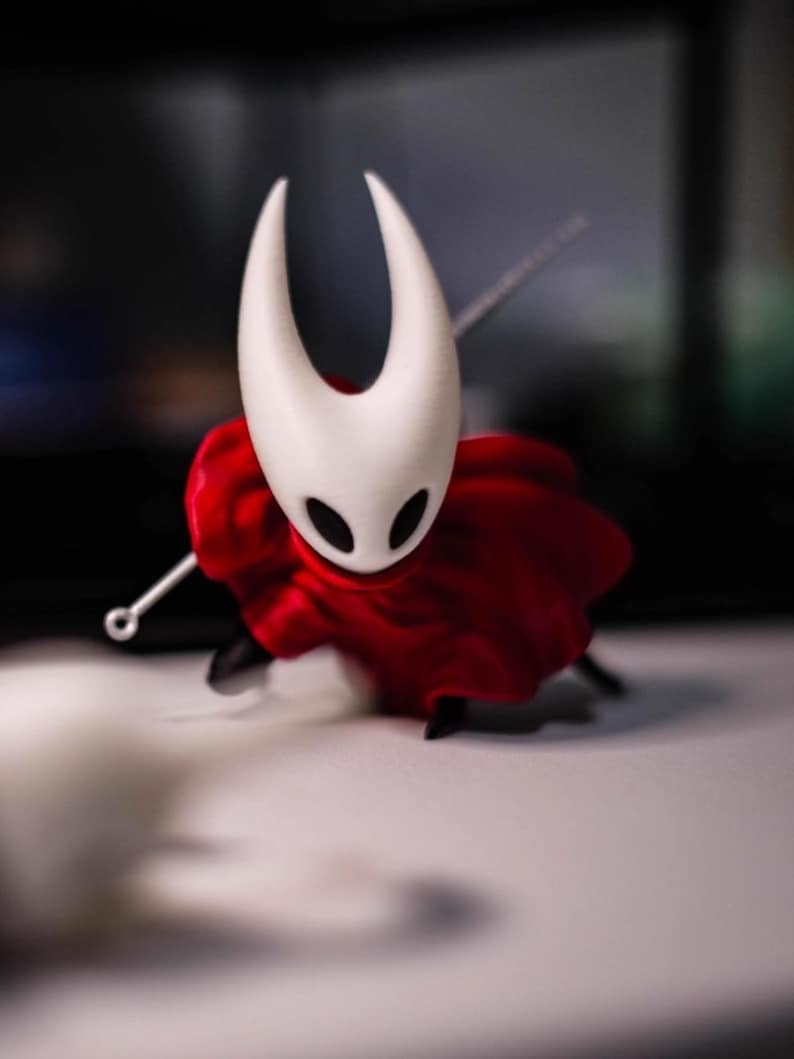 Op de afbeelding: Een beeldje van een personage uit de videogame Hollow Knight. De figuur heeft een witte kop met twee gebogen hoorns en zwarte ogen. Het draagt een rode cape en houdt een zilveren naald vast. De figuur staat op een wit oppervlak.