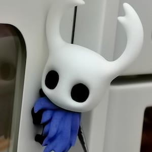 Op de afbeelding: Een wit beeldje van een personage met grote zwarte ogen en een gebogen hoofddeksel. Het personage draagt een blauwe cape en houdt een zilveren zwaard vast. Het beeldje is een verzamelobject.