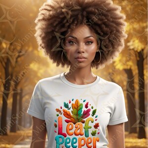 Può includere: Una donna indossa una maglietta bianca con una grafica colorata "Leaf Peeper". Il design presenta una grande foglia con accenti rossi, arancioni, verdi e blu. La donna ha i capelli ricci castani e indossa orecchini a cerchio dorati.