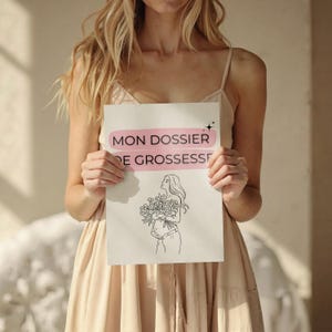 Può includere: Una persona tiene in mano un cartello bianco con la scritta "MON DOSSIER DE GROSSESSE" in rosa e nero. Sotto, un'illustrazione in bianco e nero di una persona incinta che tiene dei fiori. La persona indossa un abito colorato.