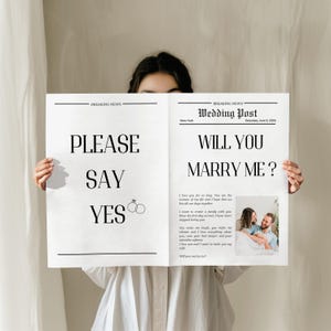 Può includere: Un cartello in stile giornale con il titolo "PLEASE SAY YES" a sinistra e "WILL YOU MARRY ME?" a destra. Il cartello include una foto di una coppia e grafiche di anelli nuziali. Lo sfondo è di colore neutro.