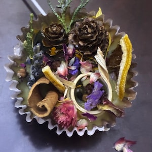 May include: A decorative candle with a light green wax base, filled with dried flowers, pine cones, and citrus peel. The candle is in a ruffled paper cup, with a variety of colors including brown, purple, and yellow.