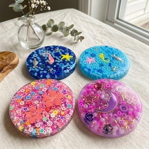 Op de afbeelding: Vier hars onderzetters met ingebedde decoratieve elementen. De onderzetters hebben blauwe, roze en paarse kleurenschema's, met ontwerpen zoals zeepaardjes, sterren, bloemen en een halve maan. Elke onderzetter heeft een diameter van ongeveer 10 cm.