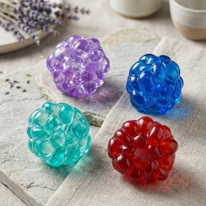Puede incluir: Cuatro bombas de baño esféricas y coloridas en tonos morado, azul, turquesa y rojo. Cada bomba de baño tiene una superficie texturizada y burbujeante. Las bombas de baño están dispuestas sobre una superficie de piedra y un paño de lino.