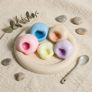 Puede incluir: Una colección de seis objetos redondos y coloridos, similares a jabones, en tonos pastel de rosa, azul, amarillo y morado, cada uno con un centro contrastante. Están dispuestos sobre un plato beige con una pequeña cuchara moteada. También hay piedras decorativas y una ramita verde.