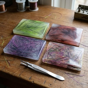 Puede incluir: Cuatro posavasos rectangulares con fibras de colores incrustadas en tonos verdes, rojos, morados y dorados. Un par de pinzas plateadas descansa sobre la superficie de madera. Los posavasos están sobre una mesa de madera.