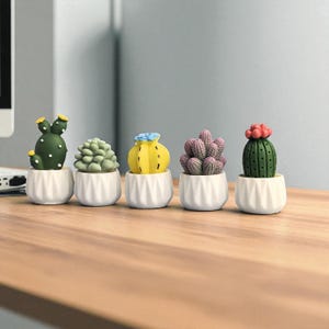 Puede incluir: Cinco pequeñas plantas de cactus en macetas de cerámica blancas. Los cactus son de varios colores, incluyendo verde, amarillo y morado, con pequeños detalles como flores amarillas y flores rojas. Las macetas tienen un diseño acanalado.