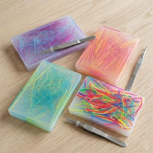 Puede incluir: Cuatro recipientes rectangulares de plástico transparente llenos de hebras de material colorido. Cada recipiente contiene una combinación de colores diferente: morado y azul, naranja y amarillo, verde y amarillo, y un arcoíris. Pinzas colocadas cerca.