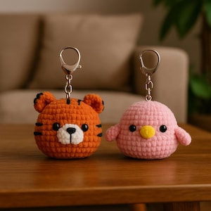 May include: Two crochet keyrings: an orange tiger with black stripes and a pink bird with a yellow beak. Both have silver key rings attached. The tiger has a white muzzle and black eyes. The bird has black eyes and small wings.