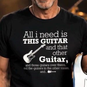 Op de afbeelding: Een zwart t-shirt met korte mouwen en witte tekst: All i need is THIS GUITAR and that other Guitar, and those guitars over there, and the guitars in the other room, and... met een kleine afbeelding van een witte elektrische gitaar.