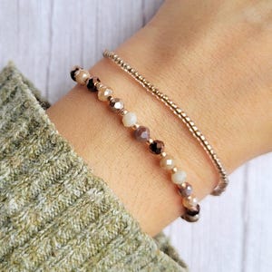 Könnte beinhalten: Ein Set aus zwei zusammen getragenen Armbändern. Ein Armband besteht aus kleinen, metallischen roségoldenen Perlen. Das zweite Armband zeigt eine Reihe facettierter, mehrfarbiger Glasperlen in Creme-, Beige-, Braun- und Roségoldtönen.