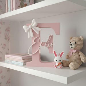 May include: A pink wooden letter "E" with a white bow and the name "Eflin" in cursive, displayed on a white shelf. A small bunny figurine and a crocheted teddy bear sit beside the letter. Books are stacked on the shelf, creating a nursery or children's room theme.
