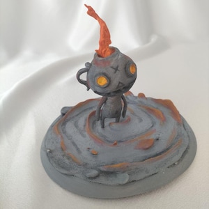 May include: A gray figurine of a small creature with orange glowing eyes and a flame-like protrusion from its head. The figure stands on a gray, circular base with orange accents, set against a white background.