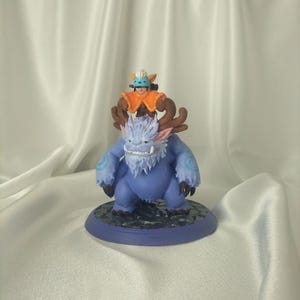 May include: A handcrafted figurine featuring a blue, furry creature with large antlers and a smaller figure riding on its back. The figures are painted with vibrant colors and detailed features, set on a circular base.