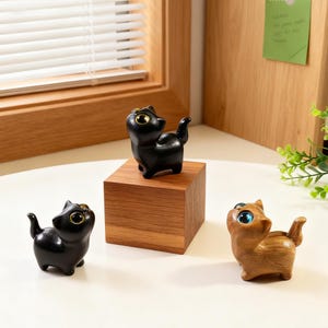 Pode incluir: Três pequenas estatuetas de gato. Duas são pretas com olhos dourados e uma é castanha com olhos azuis. Os gatos estão sobre uma superfície branca e um bloco de madeira. As estatuetas têm um acabamento brilhante e um design estilizado.