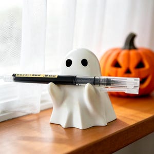 Può includere: Un portapenne a forma di fantasma in ceramica bianca con occhi neri, che tiene una penna nera con la punta trasparente. Una zucca arancione con caratteristiche intagliate è sullo sfondo. Il portapenne è su una superficie di legno.