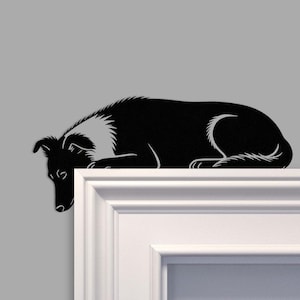 May include: A black and white silhouette of a dog lying down, with its head and front paws overhanging a white ledge. The dog has a white patch on its neck and chest. The background is a plain grey colour.