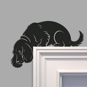 Op de afbeelding: Zwart metalen hondensilhouet, ontworpen om op een deurkozijn of plank te zitten. De hond wordt afgebeeld in een neerwaartse houding, met zijn kop en voorpoten over de rand hangend. De vacht van de hond is gedetailleerd met gebogen lijnen.