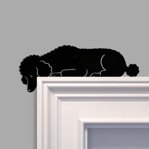 Puede incluir: Silueta negra de caniche colocada decorativamente sobre una superficie blanca. El perro está tumbado, con la cabeza y las patas delanteras colgando del borde. El fondo es gris liso.