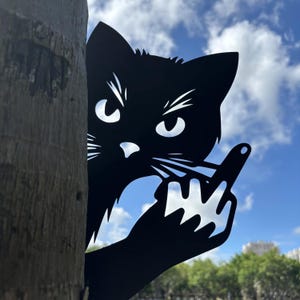 May include: Black metal cat silhouette with an angry expression, giving the middle finger. The cat is positioned behind a tree trunk, with a blue sky and clouds in the background. A humorous garden or yard decoration.
