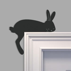 May include: A black metal rabbit silhouette decoratively placed on a white door frame. The rabbit is in a sleeping position, with its ears upright and eyes closed. The wall behind the door frame is a light gray color.