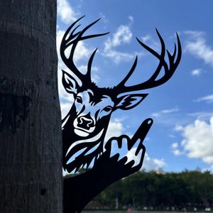 Könnte beinhalten: Schwarze Metall-Hirsch-Silhouette mit großem Geweih, die den Mittelfinger zeigt. Der Hirsch wird teilweise von einem Baumstamm verdeckt, vor einem blauen Himmel mit weißen Wolken. Ein humorvolles und freches Gartenkunstwerk.