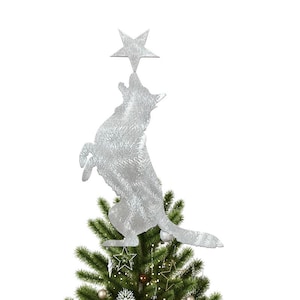 Puede incluir: Un adorno plateado para la parte superior del árbol de Navidad que representa un lobo aullando que alcanza una estrella. El lobo está colocado encima de un árbol de Navidad decorado con adornos y luces. Una decoración festiva.
